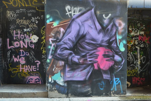 street art and graffiti in an alley, on a wall and in a doorway. On the wall is a headless man holding a pink blob in his hands, on the doorway is a lot of graffiti and tags along with the words, How long do we have?