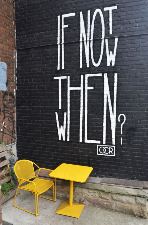 words written large in white on a black wall, if not now then when. A bright yellow table and chair sit in front of the wall. 