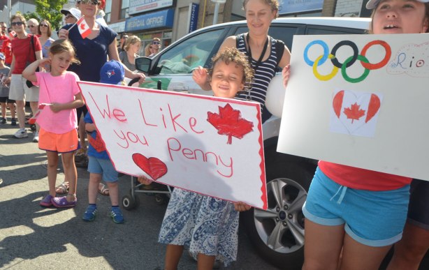 people watching a parade including a young child is holding up a red and white hand written sign that says We like you Penny. It also has a red maple leaf on it and a red heart. Another girl is holding a home made sign with the Olympics rings on it as well as a Canadian flag in a heart shape. 