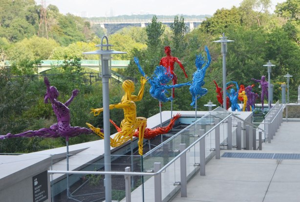 brightly coloured sculptures of figures in various active poses, made of loosely woven metal, a purple woman leaping, an orange man on his back about to catch of blue woman who is leaping head first through the air, and others as well, along the side of a building, the DVP runs beside in the background and the Bloor Viaduct bridge across the valley is in the distance