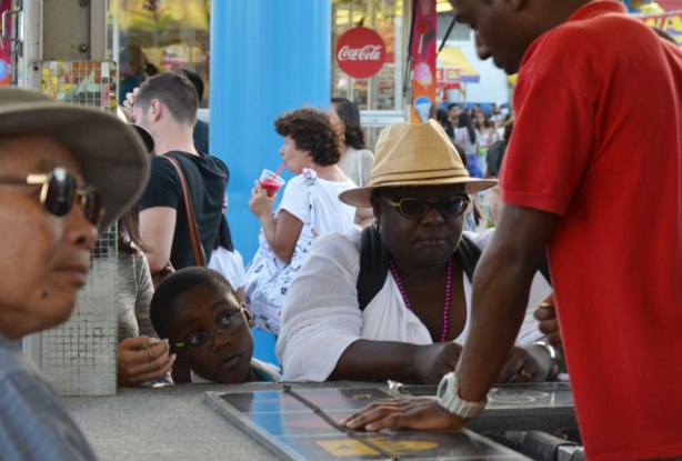 a young boy pokes his around his mother to watch her as she plays a gambling game on the midway at the Ex 