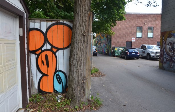 a small painting of a large orange and blue snail on a fence beside a tree in an alley 