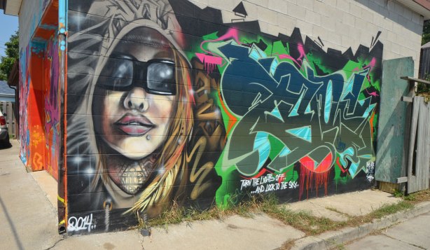 street art in an alley of a woman with long hair, wearing sunglasses, a tag beside it. 