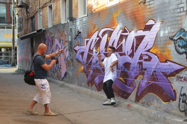making a music video in front of the street art in an alley, a young man in white t shirt is dancing to the music (and lip synching too I think) while another man, bald, is filming him and recording him. 