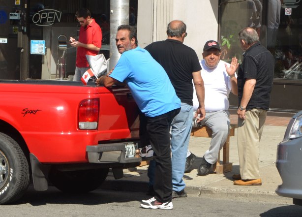 a middle age man leans against the back of a pickup truck that is parked beside a sidewalk where three older men are talking. One of them is sitting on a bench and facing the camera. 