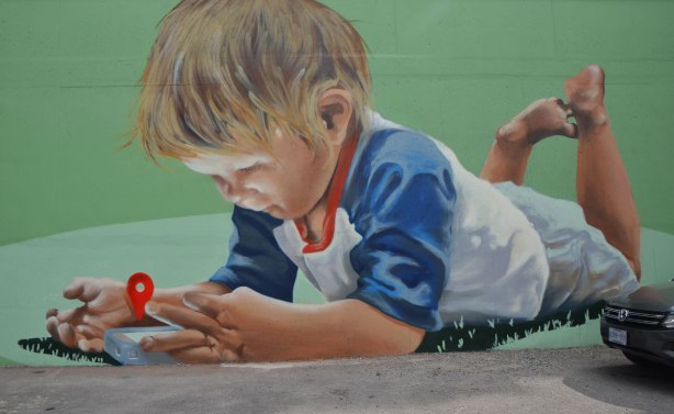 mural of a boy lying on his stomace, with his feet in the air. He islooking at an electronic device 