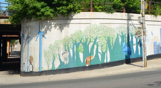 mural on a curved concrete wall, beside an intersection, showing windmills with bike parked in front, and a forest with some animals in it, fox and wolf