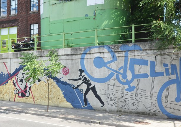 mural on the wall of an underpass, in the Junction, on Dupont, a line drawing of a railway car, with a large blue bike superimposed on top of it, a person holding a stop sign,