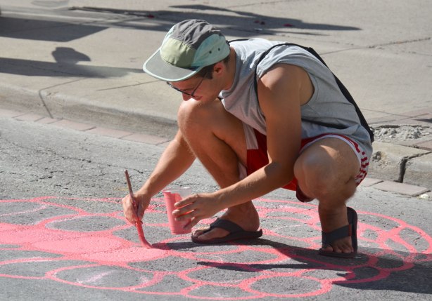 a young man paints part of a pink raspberry that is in a mural of fruits and vegetables, on a street in Kensington 