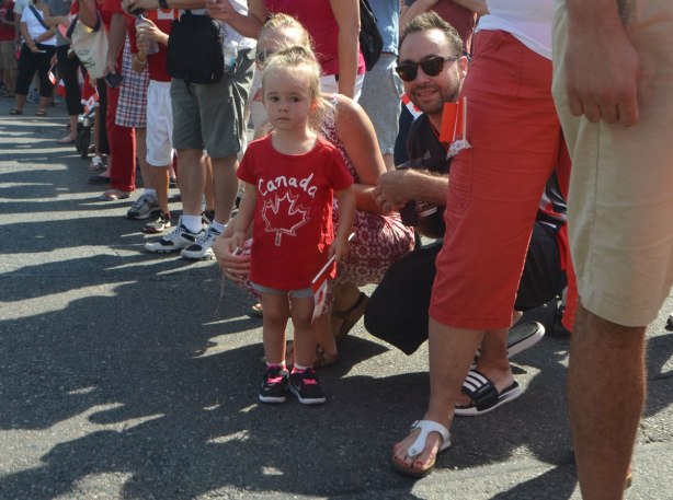a mother and father are squatting down beside his young daughter who is standing beside a street waiting for a parade to start, amongst other people, little girl is wearing a red Canada T-shirt. 