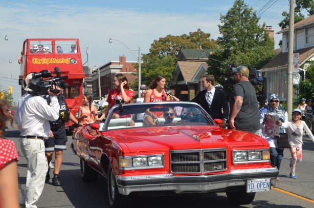 Penny Olesiak and Michelle Williams, two Canadian swimmers who won medals in swimming in the 2016 olympics, ride in the back of a red convertible, a TV camera is on one side of the car and a man interviewing Michelle is on the other side of the car. The car is moving in a parade. 