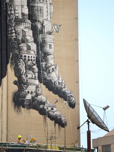 feet of a person in a mural made of paintings of trees and buildings, roots growing out of the heels. satelite dish in the mural and on the building beside the mural 