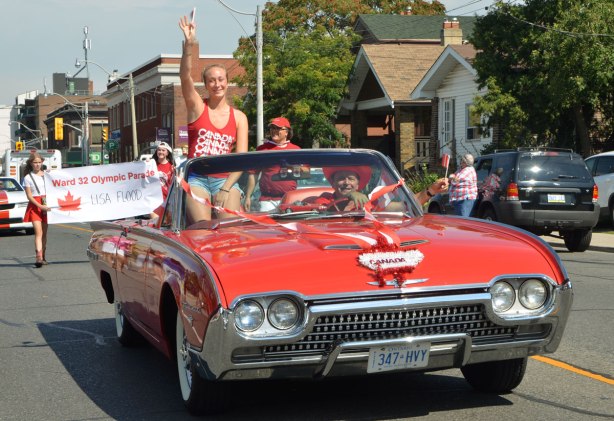 A woman Canadian athlete, Rose Cossar, rides in a red convertible in a parade. She is holding up a small Canadian flag. 