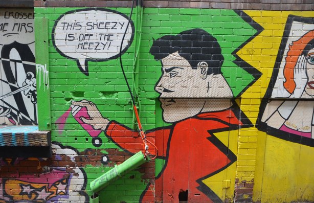 This seezy is off the heezy are the words in a word bubble coming out of the mouth of a man painted in a mural, short black hair, wearing a red shirt, and spraying a can of pink spray paint,