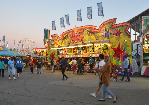 midway game, shoot out, early evening, lights on, people walking in front, ferris wheel in the distance. 