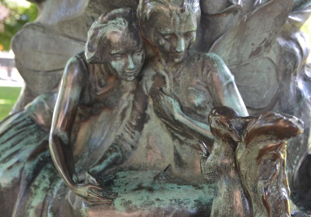 part of a sculpture, a squirrel is sitting on its back legs, looking at two fairies (women with large wings) who are looking back at the squirrel 