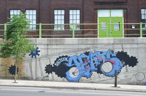 mural on a concrete wall beside a sidewalk, just before the road goes under an underpass, gears, plus a stylized industrial machine in black and blue