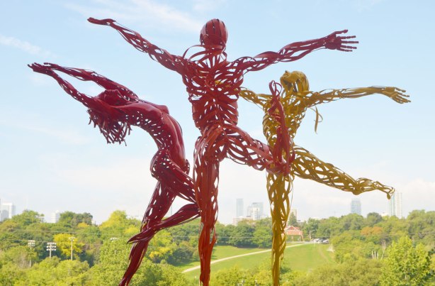 outdoor sculptures of three women dancing, one is red metal, one is orange metal and the last is yellow metal. Downtown TOronto skyline is in the distance
