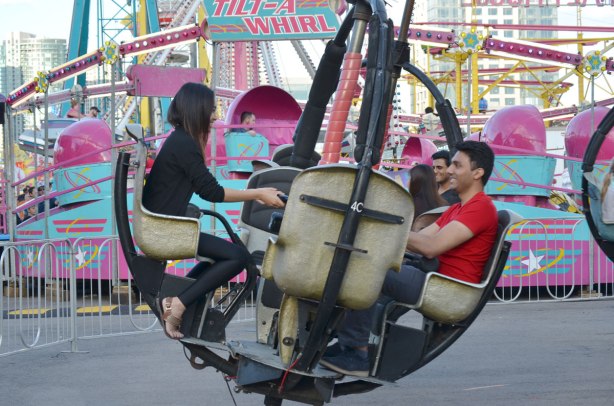 a couple ride on a midway ride at the Canadian Exhibition, behind them is the blue and pink tilt-a-whirl ride