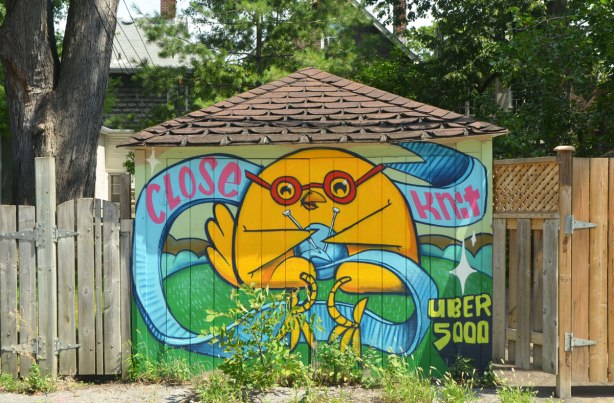 an uber 5000 art piece on a garage door, yellow uber birdie is knitting something with blue wool. the words say "close knit"