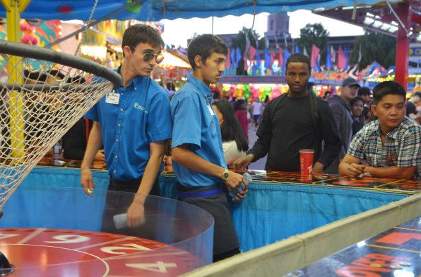 two young men in blue shirts working at one of the midway gambling games, with another man standing behind the counter