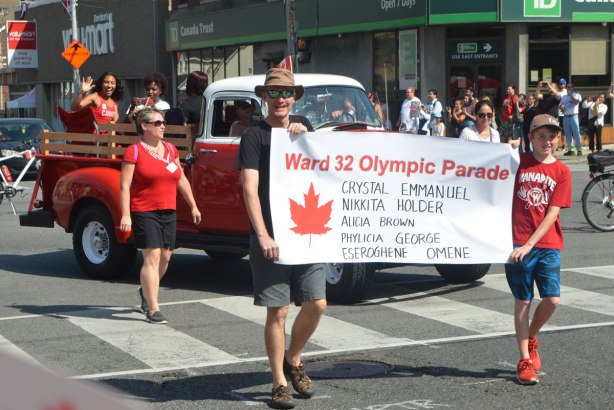 a man and a boy hold a banner in a parade, banner says ward 32 olympic parade. And then it lists the athletes that are riding in the old red and white pick-up truck behind the banner, Crystal Emmanuel, Alicia Brown, Nikkita Holder, Philicia George and Eseroghene Omene 