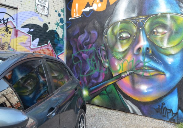 mural of a large face with paint brush by the mouth, wearing glasses, mural seems to be looking at a car that is parked in front of it, with reflections of the mural in the car window