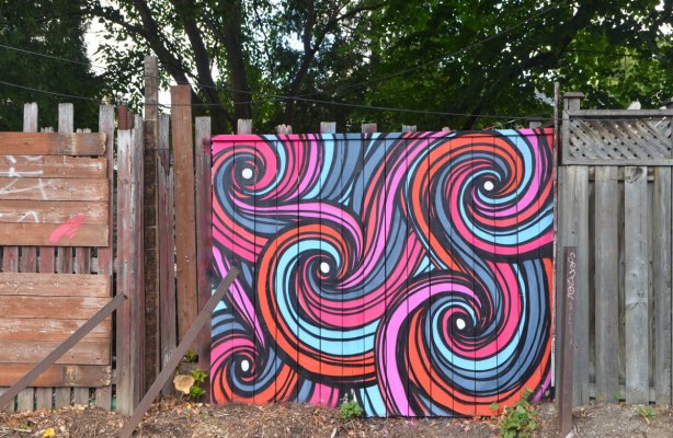 a section of wooden fence covered with street art in pink and orange and turquoise and purple striped swirls 