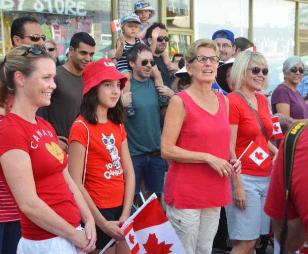 Ontario premier Kathleen Wynne stands amongst some people on a sidewalk who are waiting for a parade to start