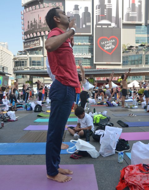 a man in red shirt and blue pants is standing tall, others around him also standing in yoga position, a boy is sitting on the ground with his legs crossed and looking at something in his lap 