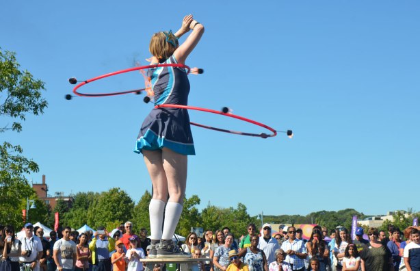 woman standing on a stool with two hula hoops around her middle, both of which have 5 or 6 spots that are on fire, in front of a crowd of people sitting on the grass