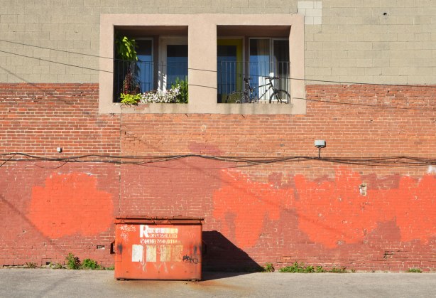 beige wall on top, rust coloured wall below, with orange splotches. Orange metal trash bin on ground, two window in upper part, both recessed. One with a bike and one with flower pots. 