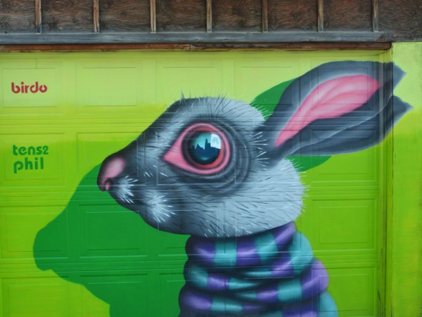 birdo street art of a rabbit head on a bright green garage door, grey head, pink inside of the long ears, purple and turquoise striped neck. 