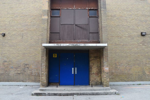 blue double doors in a drab brown brick building, with brown metal inserts covering what was once a large window above the door. Two small windows remain. 