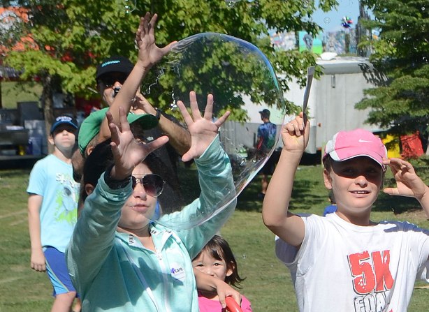 very large bubbles being made in front of a crowd of children and adults, kids chasing and trying to catch and burst the bubbles, just at the moment that the bubble breaks