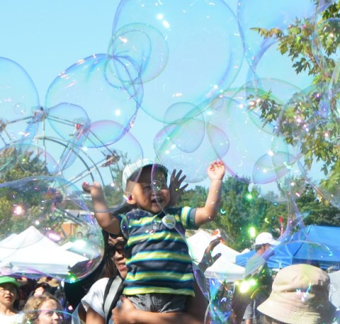 very large bubbles being made in front of a crowd of children and adults, kids chasing and trying to catch and burst the bubbles, young boy being held up by his mother to reach the bubbles