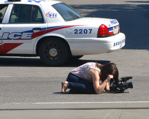 a camera woman has her camera on the ground and she is kneeling behind it and trying to take a picture. She is bare footed. There is a police car parked behind her. She is on the street, photographing a parade