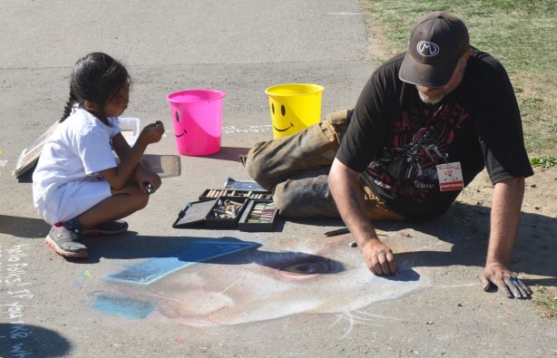 man is drawing a picture with chalk on the sidewalk. A little girl is squatting beside him and watching him work 