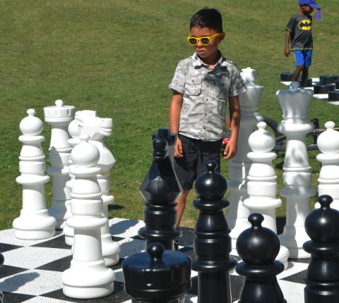 a young boy with bright yellow rimmed sunglasses stands on a large chess board contemplating his next move in the game of chess. 