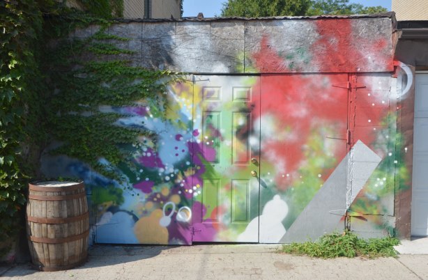 abstract multicoloured street art on a wall and door, part of wall is also covered with ivy. old wooden barrel sits by wall 