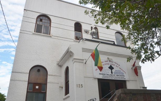 front of a white building, two storeys, with round top windows, two flags flying by the door. Sign says Debre Selam St. Michael Ethiopian Orthodox Tewahedo Church. Small cross above the entranceway