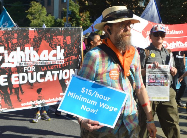 a man with a large beard, wearing a straw hat and an orange bandana around his neck, carries a sign that says $15/hour minimum wage now, in a parade. behind him is a banner protesting the cost of university education and demanding free university education