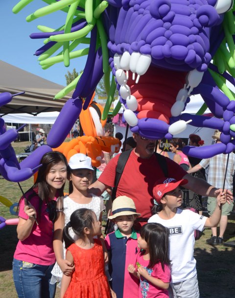 a large monster made of purple, red and green balloons is manned by a man, an Asian family is having their picture taken in front of the monster