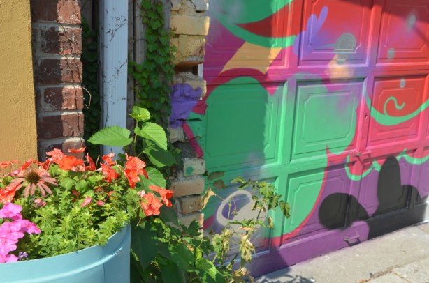 an old blue plastic barrel that has been turned into a planter, with pink flowers and greenery, in front of a garage door that has been painted in abstract street art in bright colours. 