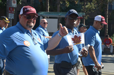 men in light blue T-shirts and wearing baseball caps, pass by in a parade, one is holding a small union flag (amalgamated transit union) and one is giving a thumbs up 