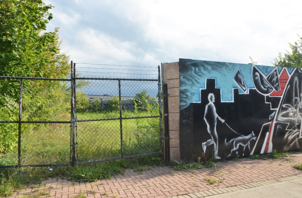 chain link fence with barbed wire above it on the left side and then a wood fence on the right, wood fence has been painted with a mural and here in the mural is a man walking a dog past a building