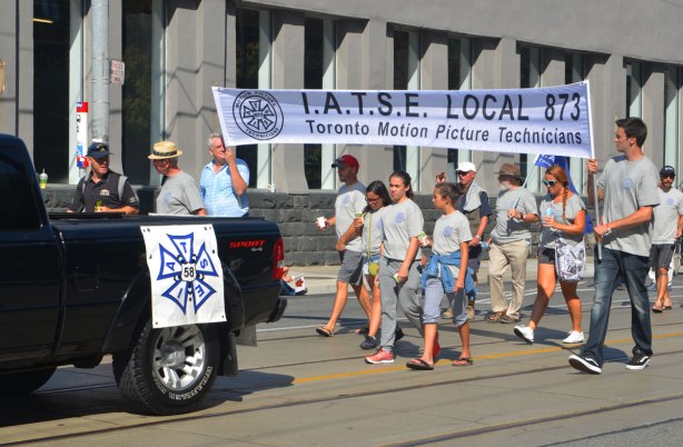 a group of people in grey t-shirts walk behind a black pick up truck in a parade, carrying a banner that says IATSE LOcal 873 union, Toronto Motion Picture Technicians. 
