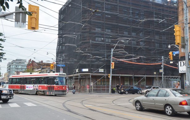 intersection, TTC street car turning left, a grey car near the intersection, a few pedestrians, a large building wrapped in black netting as the building is being cleaned and renovated. 