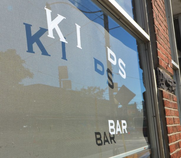 window of a coffee shop where some of the letters have been removed. It now says Kids Bar. Shadows of the letters are on the blind that covers the inside of the window of the now closed shop