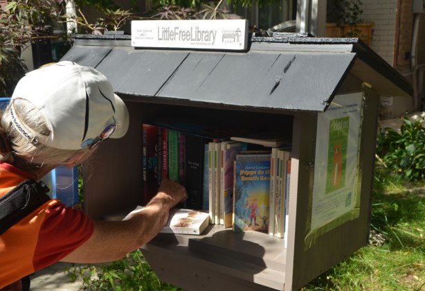 a woman in a white baseball cap is reaching into a 'little free library' shelf of books outside a house 